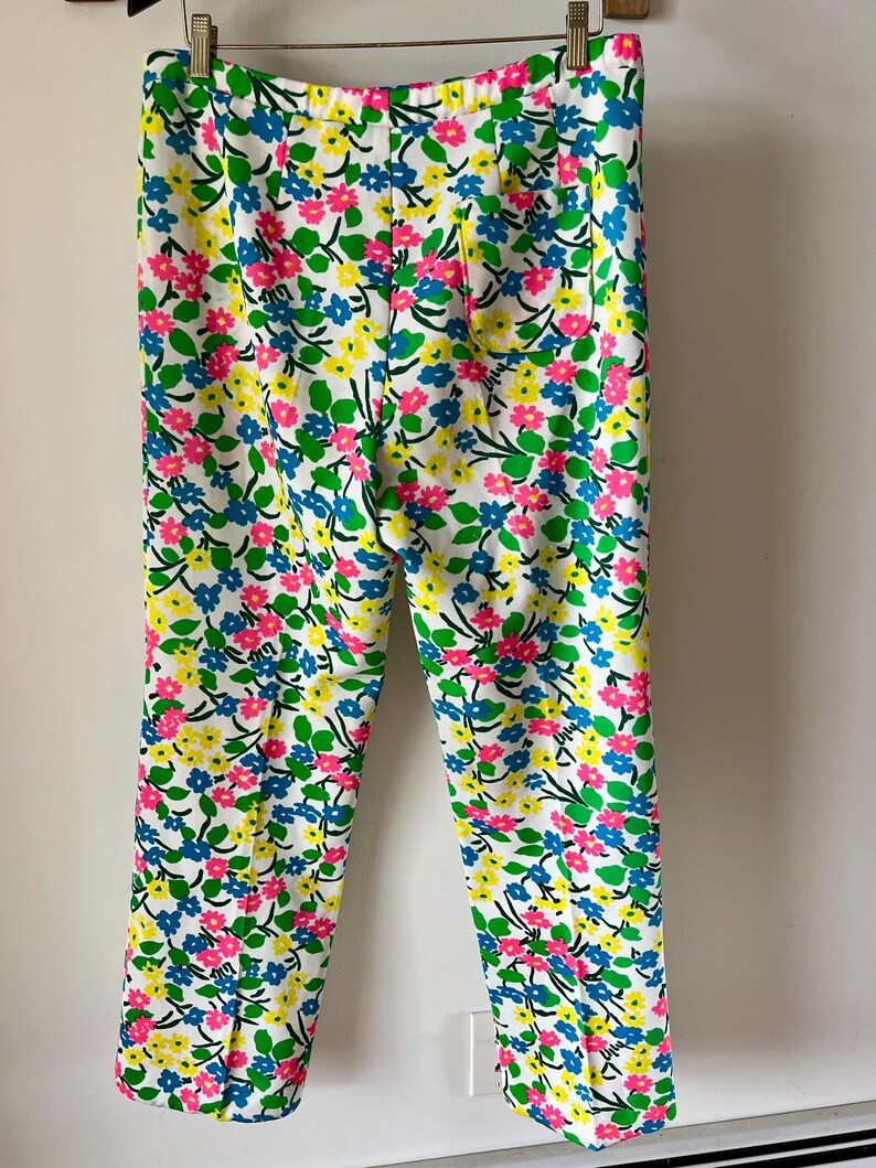 Puede incluir: Pantalones blancos con un estampado floral vibrante con flores rosas, azules y amarillas con hojas verdes. Los pantalones son de corte recto y cuelgan de una percha.