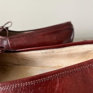Pode incluir: Close-up de um par de sapatilhas de bailarina em couro bord&ocirc;. Os sapatos t&ecirc;m bico arredondado e um pequeno detalhe de la&ccedil;o. O interior do sapato &eacute; de cor bege claro. O tamanho &eacute; indicado como "0029-7-39".