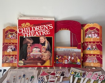 Pode incluir: Um conjunto de jogo Children's Theatre com uma caixa vermelha com o texto "Award Winning Children's Theatre", um palco de cartão dobrável com cortinas vermelhas e várias figuras de papel em paus para contar histórias.