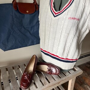 Pode incluir: Um colete branco de malha canelada com listras vermelhas e azuis, bordado com "STANFORD" em vermelho. Uma sacola azul marinho com al&ccedil;as de couro marrom e um par de mocassins de couro bord&ocirc; tamb&eacute;m s&atilde;o vis&iacute;veis.