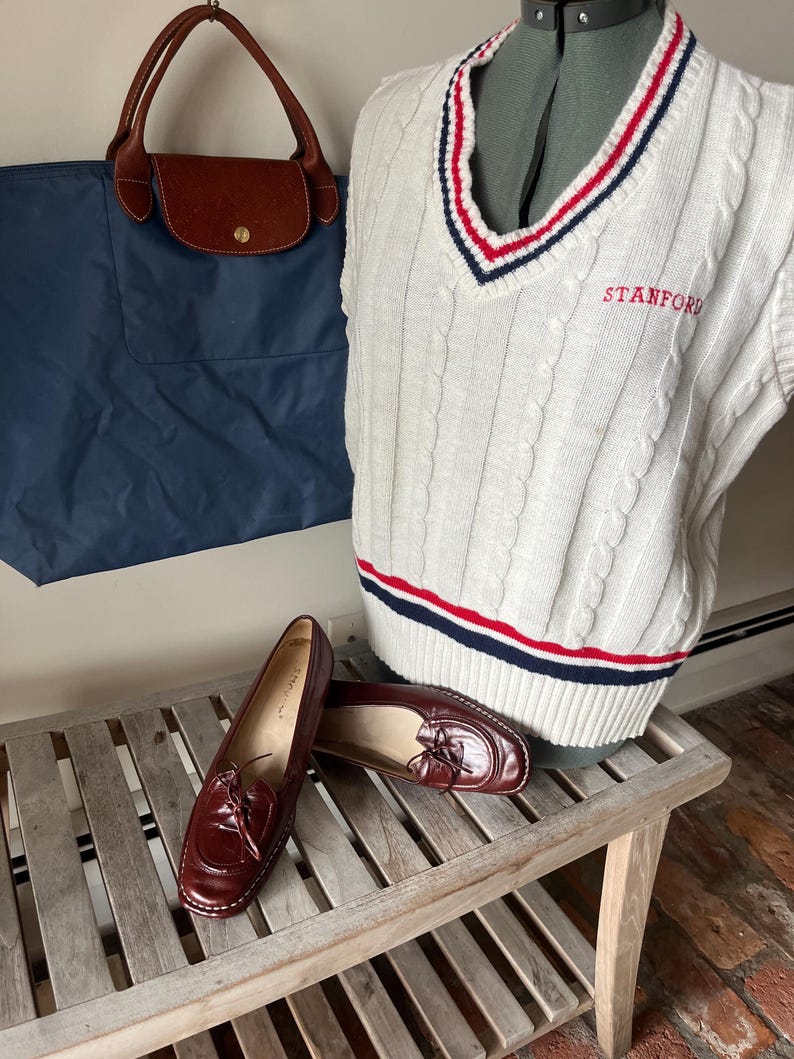 Pode incluir: Um colete branco de malha canelada com listras vermelhas e azuis, bordado com "STANFORD" em vermelho. Uma sacola azul marinho com al&ccedil;as de couro marrom e um par de mocassins de couro bord&ocirc; tamb&eacute;m s&atilde;o vis&iacute;veis.