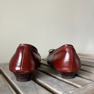 Pode incluir: Um par de sapatilhas vintage em couro bord&ocirc;. Os sapatos t&ecirc;m bico redondo, salto pequeno e detalhe de la&ccedil;o na parte superior. O couro parece bem conservado, com um ligeiro brilho. Os sapatos est&atilde;o sobre uma superf&iacute;cie de madeira.