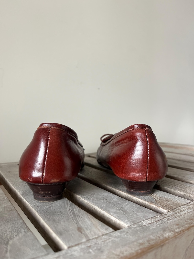 Pode incluir: Um par de sapatilhas vintage em couro bord&ocirc;. Os sapatos t&ecirc;m bico redondo, salto pequeno e detalhe de la&ccedil;o na parte superior. O couro parece bem conservado, com um ligeiro brilho. Os sapatos est&atilde;o sobre uma superf&iacute;cie de madeira.