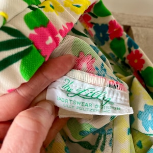 Puede incluir: Primer plano de una tela con estampado floral con flores rosas, amarillas, azules y verdes sobre un fondo blanco. La etiqueta dice "The Lilly Sportswear Division Lilly Pulitzer Inc."