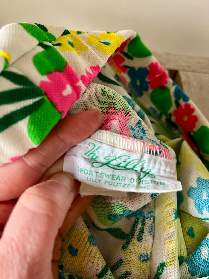 Puede incluir: Primer plano de una tela con estampado floral con flores rosas, amarillas, azules y verdes sobre un fondo blanco. La etiqueta dice "The Lilly Sportswear Division Lilly Pulitzer Inc."