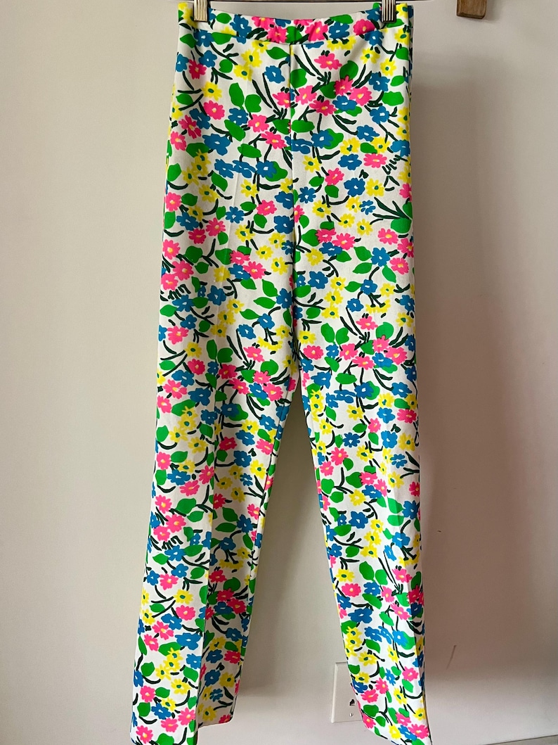 Puede incluir: Pantalones blancos con un vibrante estampado floral. El dise&ntilde;o presenta flores rosas, azules y amarillas con hojas verdes sobre un fondo blanco. Los pantalones est&aacute;n colgados en una percha de madera.