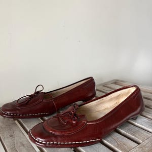 Pode incluir: Um par de mocassins de couro bord&ocirc; com bico redondo e salto pequeno. Os sapatos apresentam um adorno de borla e costuras brancas ao redor da sola. Os mocassins s&atilde;o exibidos em uma superf&iacute;cie de madeira.