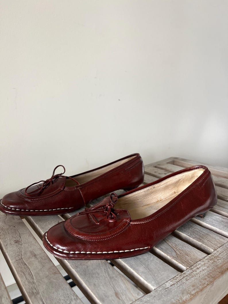 Pode incluir: Um par de mocassins de couro bord&ocirc; com bico redondo e salto pequeno. Os sapatos apresentam um adorno de borla e costuras brancas ao redor da sola. Os mocassins s&atilde;o exibidos em uma superf&iacute;cie de madeira.