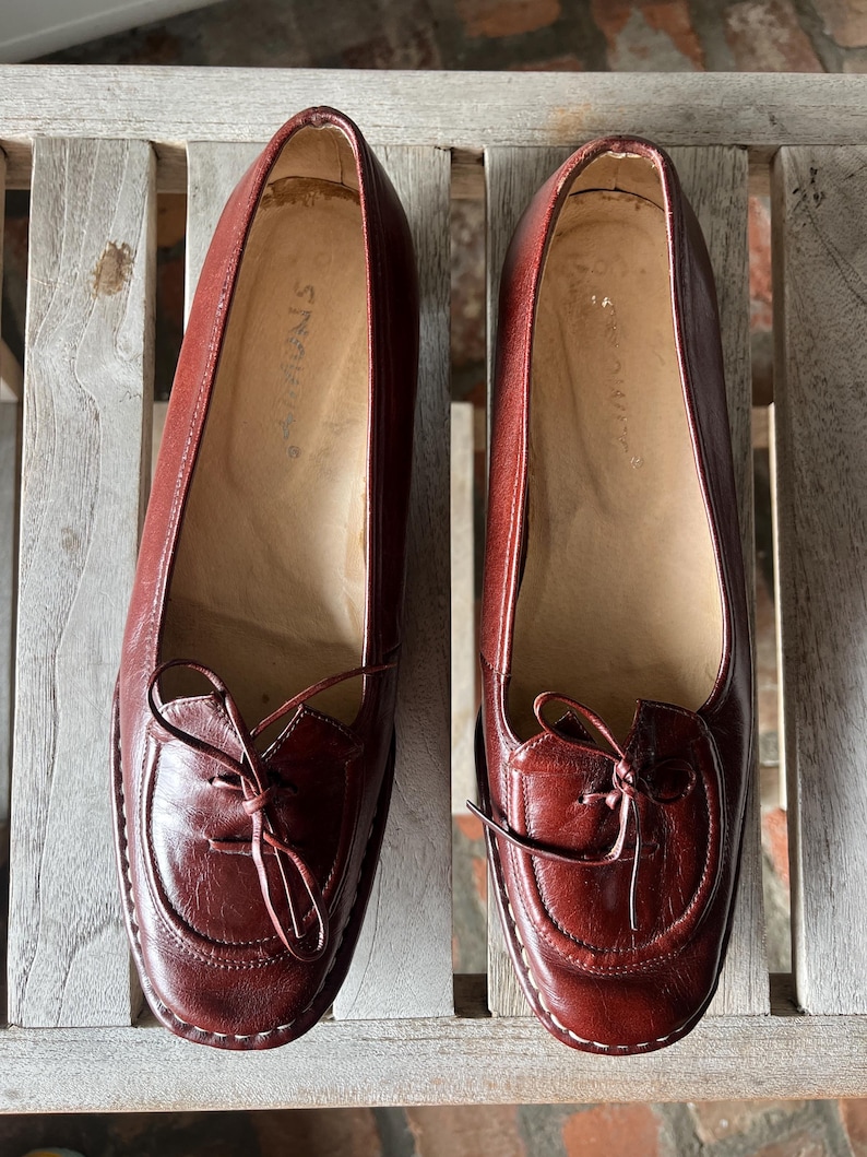Pode incluir: Um par de sapatilhas de couro bord&ocirc; com bico redondo e la&ccedil;o decorativo. Os sapatos t&ecirc;m um design costurado nas bordas e s&atilde;o exibidos em uma superf&iacute;cie de madeira. As sapatilhas s&atilde;o um estilo cl&aacute;ssico.