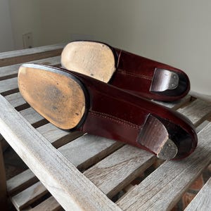Pode incluir: Um par de sapatos de couro bord&ocirc; escuro, provavelmente sapatos sociais, com saltos de madeira e solas gastas. Os sapatos est&atilde;o posicionados sobre uma superf&iacute;cie de ripas de madeira desgastada, mostrando sua apar&ecirc;ncia envelhecida e artesanato.