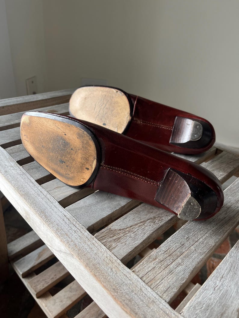 Pode incluir: Um par de sapatos de couro bord&ocirc; escuro, provavelmente sapatos sociais, com saltos de madeira e solas gastas. Os sapatos est&atilde;o posicionados sobre uma superf&iacute;cie de ripas de madeira desgastada, mostrando sua apar&ecirc;ncia envelhecida e artesanato.