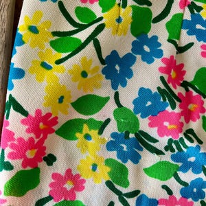 Puede incluir: Primer plano de una tela con estampado floral. El dise&ntilde;o presenta flores rosas, amarillas y azules con hojas y tallos verdes sobre un fondo blanco. La tela tiene un aspecto texturizado.