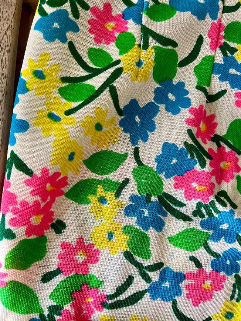 Puede incluir: Primer plano de una tela con estampado floral. El dise&ntilde;o presenta flores rosas, amarillas y azules con hojas y tallos verdes sobre un fondo blanco. La tela tiene un aspecto texturizado.