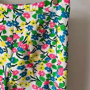 Puede incluir: Primer plano de unos pantalones blancos con un vibrante estampado floral. El dise&ntilde;o presenta flores rosas, azules y amarillas con hojas verdes, sobre un fondo blanco. Se puede ver un peque&ntilde;o bolsillo.