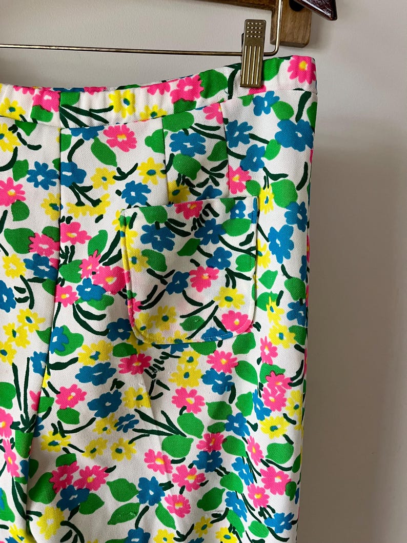 Puede incluir: Primer plano de unos pantalones blancos con un vibrante estampado floral. El dise&ntilde;o presenta flores rosas, azules y amarillas con hojas verdes, sobre un fondo blanco. Se puede ver un peque&ntilde;o bolsillo.