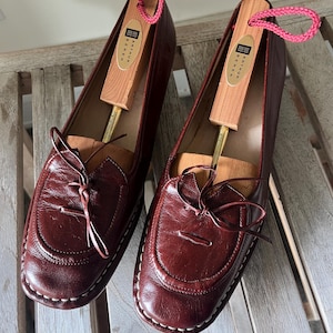 Pode incluir: Um par de mocassins de couro bord&ocirc; com costuras brancas e um la&ccedil;o decorativo. Cada sapato cont&eacute;m uma forma de sapato de madeira com um la&ccedil;o de cord&atilde;o rosa. Os sapatos s&atilde;o exibidos em uma superf&iacute;cie de madeira.