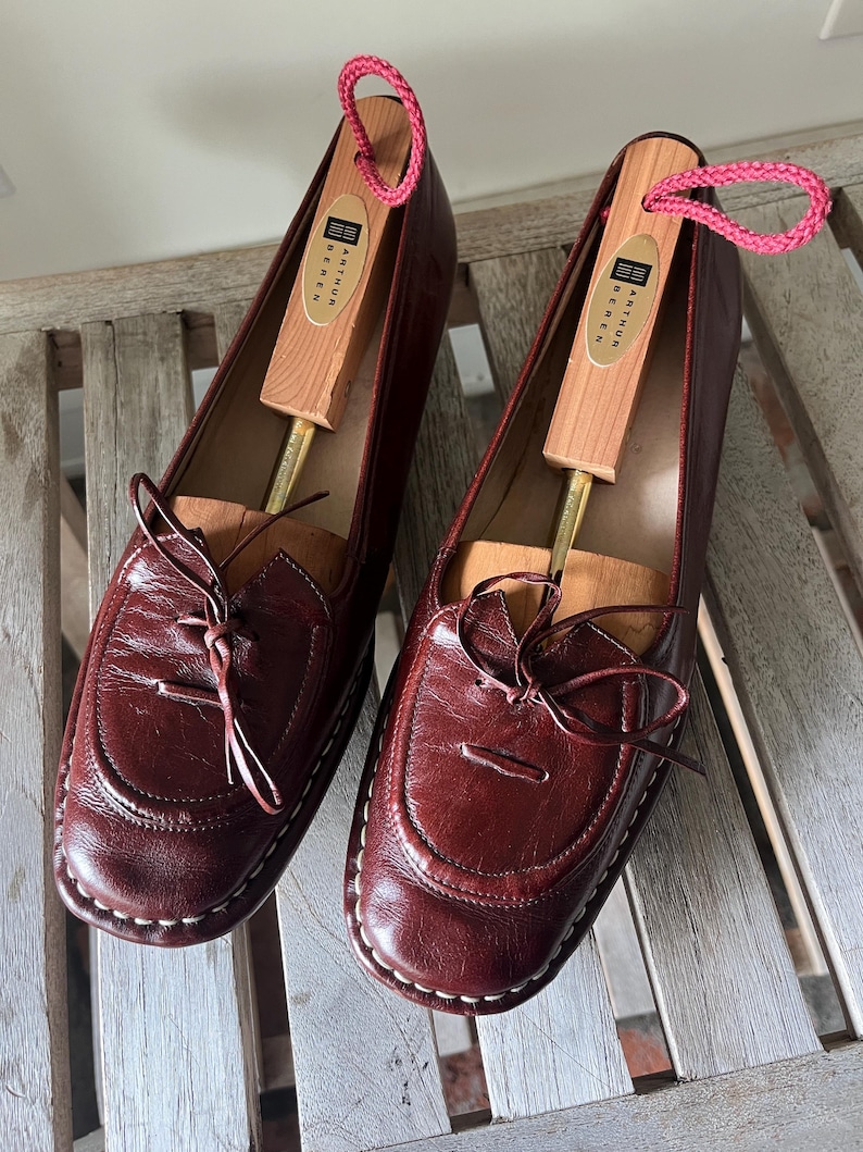 Pode incluir: Um par de mocassins de couro bord&ocirc; com costuras brancas e um la&ccedil;o decorativo. Cada sapato cont&eacute;m uma forma de sapato de madeira com um la&ccedil;o de cord&atilde;o rosa. Os sapatos s&atilde;o exibidos em uma superf&iacute;cie de madeira.