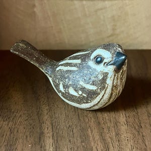 May include: A small ceramic bird figurine featuring a speckled brown and cream glaze. The bird has a dark, pointed beak, a round body with textured wing details, and a short tail. The surface has a matte, earthy finish with visible brushstrokes.