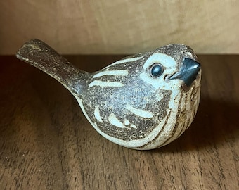 Pode incluir: Uma pequena estatueta de pássaro em cerâmica com um esmalte salpicado em castanho e creme. O pássaro tem um bico escuro e pontiagudo, um corpo redondo com detalhes de asas texturizados e uma cauda curta. A superfície tem um acabamento mate.