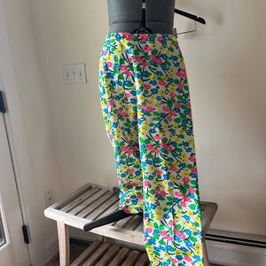 Puede incluir: Pantalones blancos con un vibrante estampado floral de flores rosas, azules y amarillas. Los pantalones se exhiben en un maniqu&iacute;, mostrando su dise&ntilde;o de pierna ancha. El estampado floral se encuentra sobre un fondo blanco.