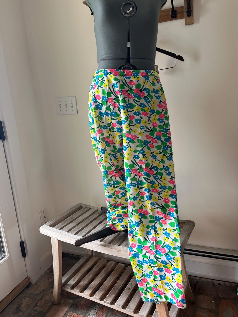 Puede incluir: Pantalones blancos con un vibrante estampado floral de flores rosas, azules y amarillas. Los pantalones se exhiben en un maniqu&iacute;, mostrando su dise&ntilde;o de pierna ancha. El estampado floral se encuentra sobre un fondo blanco.