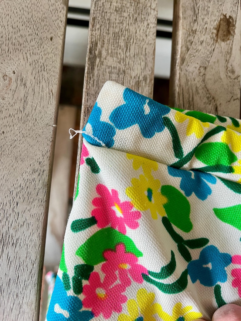 Puede incluir: Primer plano de una tela con estampado floral. La tela presenta un fondo blanco con flores rosas, amarillas y azules brillantes, junto con hojas verdes. La tela es ligeramente texturizada y parece ser parte de una prenda.