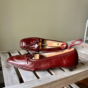 Pode incluir: Um par de sapatilhas de couro bord&ocirc; com um pequeno salto e detalhe de la&ccedil;o. Os sapatos est&atilde;o numa prateleira de madeira, com uma forma de sapato em cada um. Costuras brancas acentuam as bordas.