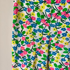 Puede incluir: Pantalones blancos con un vibrante estampado floral con flores rosas, amarillas y azules, acentuadas por hojas verdes. Los pantalones est&aacute;n colgados en una percha de metal, mostrando el dise&ntilde;o colorido.