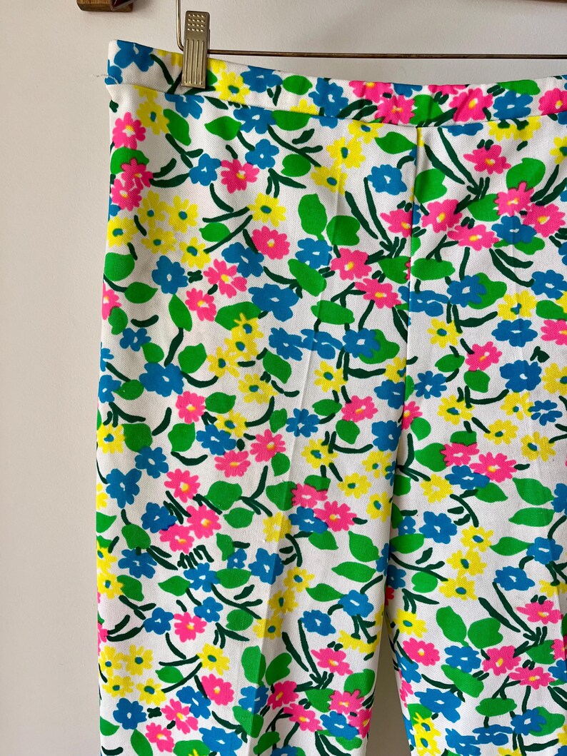 Puede incluir: Pantalones blancos con un vibrante estampado floral con flores rosas, amarillas y azules, acentuadas por hojas verdes. Los pantalones est&aacute;n colgados en una percha de metal, mostrando el dise&ntilde;o colorido.
