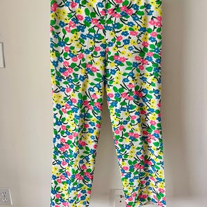 Puede incluir: Pantalones blancos con un estampado floral vibrante. El dise&ntilde;o presenta flores rosas, azules y amarillas con hojas verdes dispersas por la tela. Los pantalones cuelgan de una percha de madera.