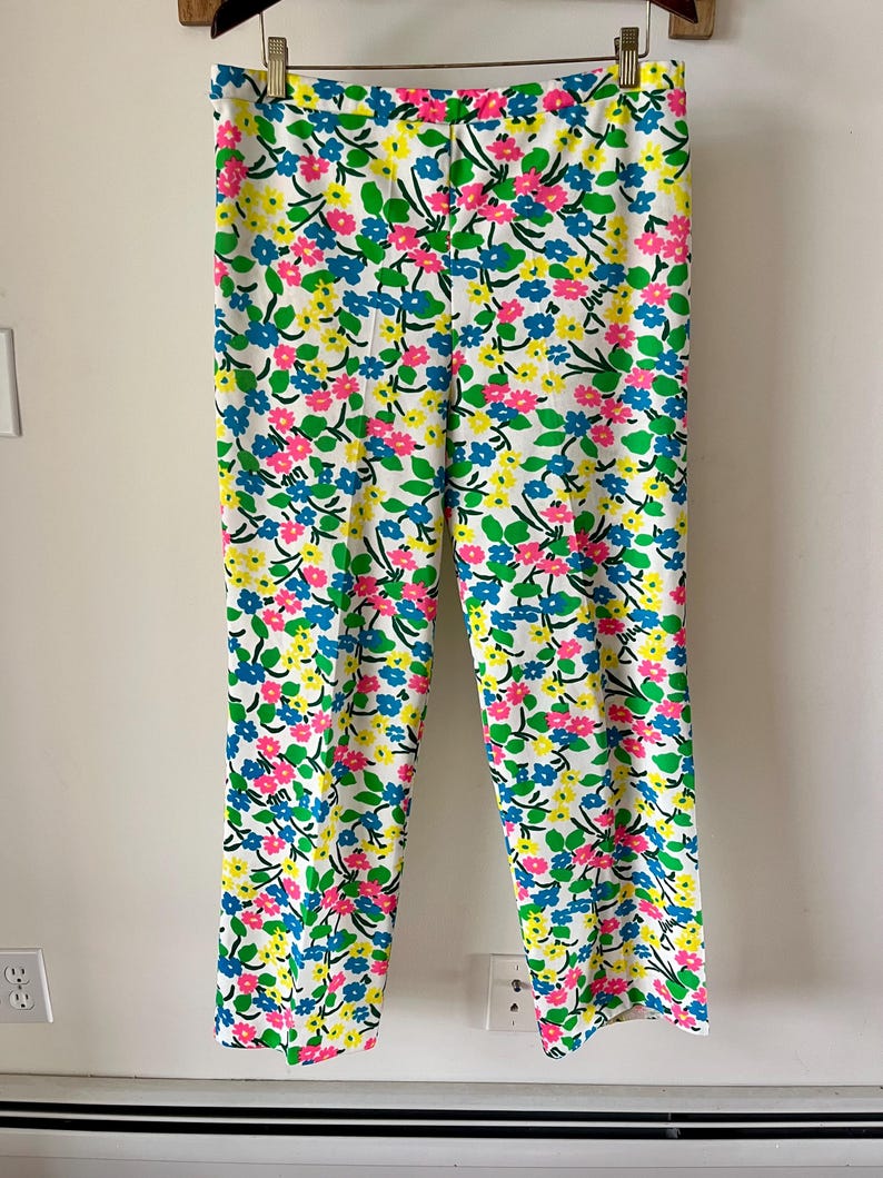 Puede incluir: Pantalones blancos con un estampado floral vibrante. El dise&ntilde;o presenta flores rosas, azules y amarillas con hojas verdes dispersas por la tela. Los pantalones cuelgan de una percha de madera.