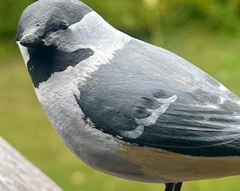Pode incluir: Uma estatueta de pássaro de madeira pintada à mão com uma carapuça e garganta pretas, uma mancha branca na bochecha e um dorso cinzento com marcas brancas nas asas. O pássaro tem a parte inferior castanha e apoia-se em finas pernas de metal preto.