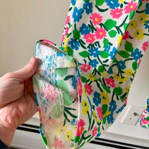 Puede incluir: Primer plano de una tela con estampado floral sobre fondo blanco, adornada con flores azules, rosas y amarillas. Se aprecia el pu&ntilde;o de la prenda, con un ribete de encaje con motivos florales rosas y azules.