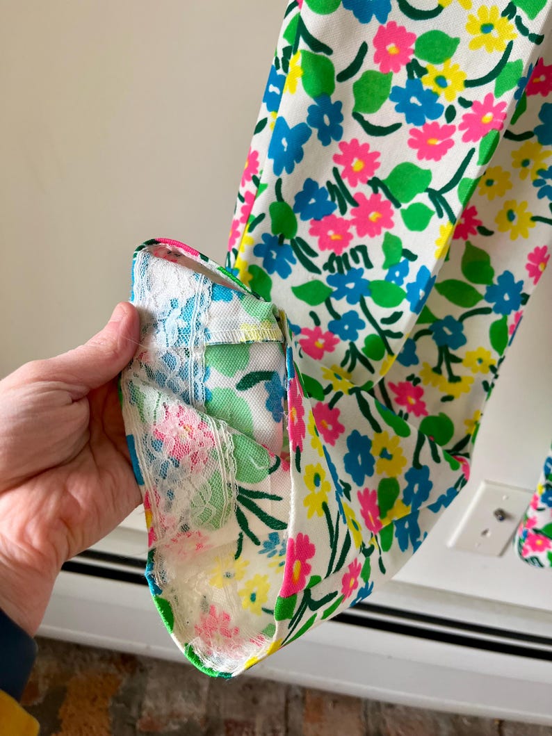 Puede incluir: Primer plano de una tela con estampado floral sobre fondo blanco, adornada con flores azules, rosas y amarillas. Se aprecia el pu&ntilde;o de la prenda, con un ribete de encaje con motivos florales rosas y azules.