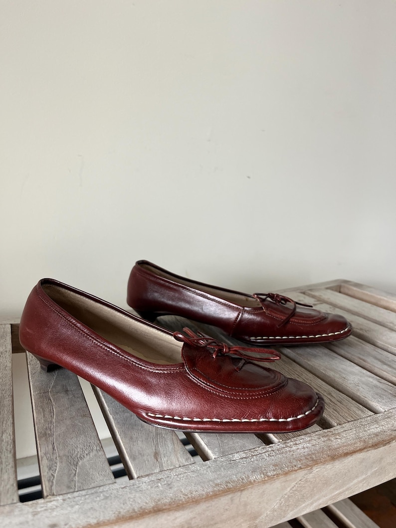 Pode incluir: Um par de mocassins de couro bord&ocirc; com saltos pequenos. Os sapatos t&ecirc;m bico arredondado, costura decorativa e um pequeno detalhe de la&ccedil;o na g&aacute;spea. S&atilde;o exibidos em uma superf&iacute;cie de madeira.