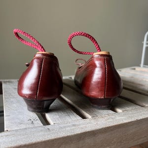 Pode incluir: Um par de sapatilhas de bailarina vintage em couro castanho com saltos pequenos. Cada sapato tem um la&ccedil;o de cord&atilde;o rosa inserido no calcanhar. Os sapatos s&atilde;o exibidos numa superf&iacute;cie de ripas de madeira.