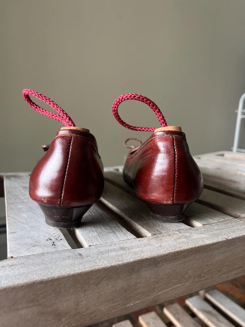 Pode incluir: Um par de sapatilhas de bailarina vintage em couro castanho com saltos pequenos. Cada sapato tem um la&ccedil;o de cord&atilde;o rosa inserido no calcanhar. Os sapatos s&atilde;o exibidos numa superf&iacute;cie de ripas de madeira.