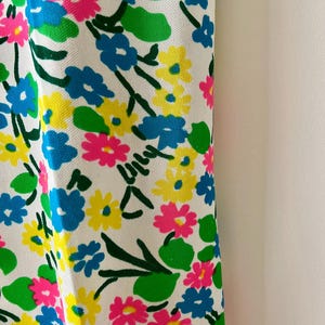Puede incluir: Primer plano de una tela blanca con un vibrante estampado floral. El dise&ntilde;o presenta grupos de flores rosas, azules y amarillas con hojas y tallos verdes. Las flores est&aacute;n dispersas por la tela, creando un dise&ntilde;o alegre y colorido.