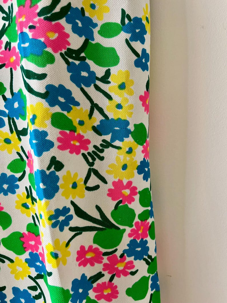 Puede incluir: Primer plano de una tela blanca con un vibrante estampado floral. El dise&ntilde;o presenta grupos de flores rosas, azules y amarillas con hojas y tallos verdes. Las flores est&aacute;n dispersas por la tela, creando un dise&ntilde;o alegre y colorido.