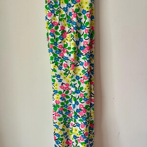 Puede incluir: Pantalones blancos con un vibrante estampado floral con flores rosas, azules y amarillas. Las flores se colocan sobre un fondo blanco con hojas verdes y tallos negros. Los pantalones est&aacute;n colgados de una percha met&aacute;lica.