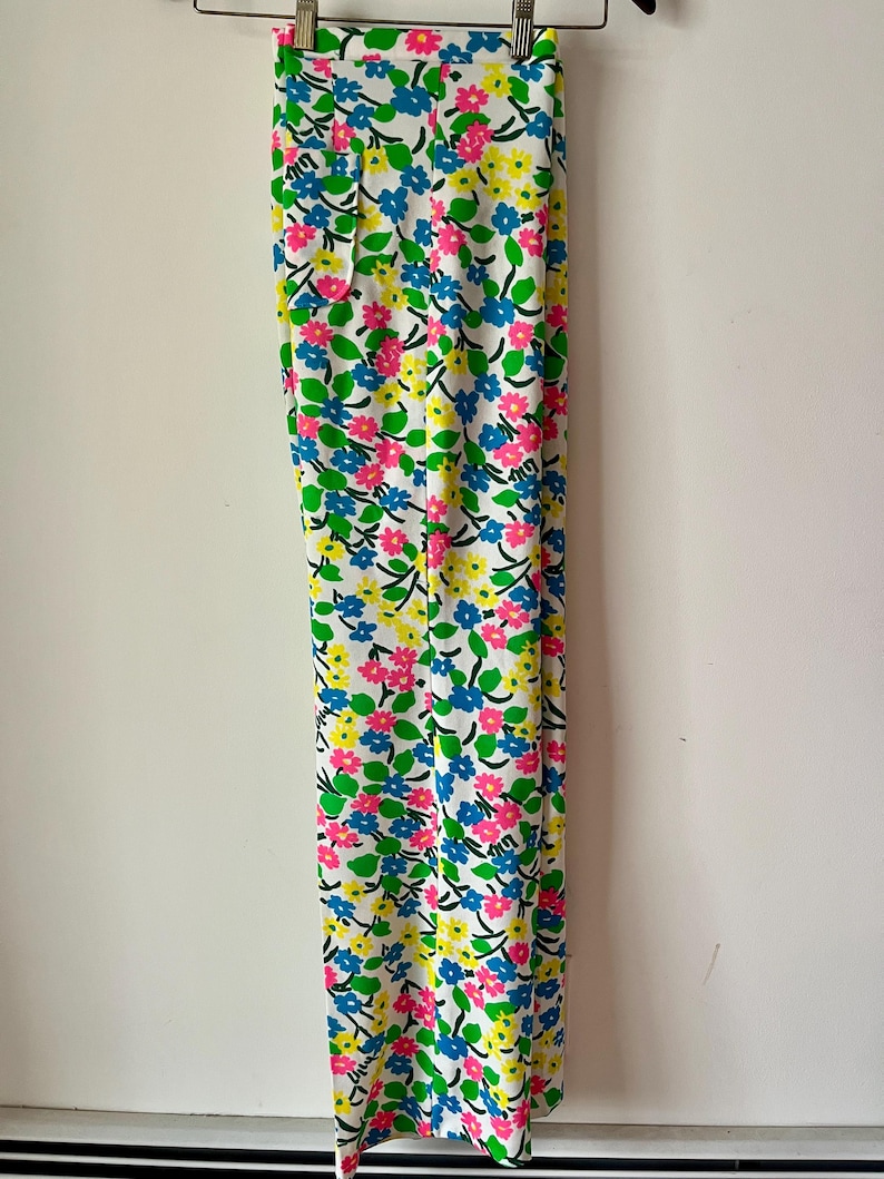 Puede incluir: Pantalones blancos con un vibrante estampado floral con flores rosas, azules y amarillas. Las flores se colocan sobre un fondo blanco con hojas verdes y tallos negros. Los pantalones est&aacute;n colgados de una percha met&aacute;lica.