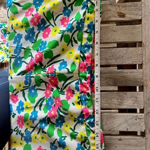 Puede incluir: Una tela blanca con un estampado floral en rosa, azul y amarillo. La tela mide aproximadamente 43 cm de largo. Hojas y tallos verdes se intercalan entre las flores. Una cinta m&eacute;trica se coloca a lo largo del borde de la tela.