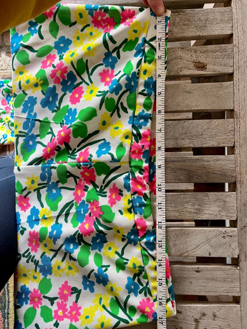 Puede incluir: Una tela blanca con un estampado floral en rosa, azul y amarillo. La tela mide aproximadamente 43 cm de largo. Hojas y tallos verdes se intercalan entre las flores. Una cinta m&eacute;trica se coloca a lo largo del borde de la tela.