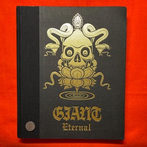 以下が含まれることがあります： 蛇と蓮の花が描かれた金色のイラストが施された黒いハードカバーの本。表紙には金色の文字で「Giant Eternal」というタイトルが書かれている。