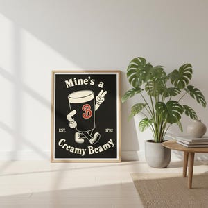 Op de afbeelding: Een ingelijste poster met een zwarte achtergrond toont een cartoon bierpul met armen en benen, die een vredesteken geeft. De tekst luidt "Mine's a Creamy Beamy" met "Est. 1792" eronder. De poster staat in een kamer met een plant.