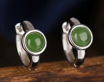 May include: A pair of silver hoop earrings, each featuring a round, vibrant green gemstone. The earrings are displayed on a wooden surface, with a dark blue background.