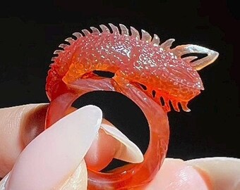 May include: A translucent orange ring featuring a detailed lizard design. The ring showcases the lizard's head and spine, with a smooth band. The ring is held against a dark background, highlighting its intricate details.