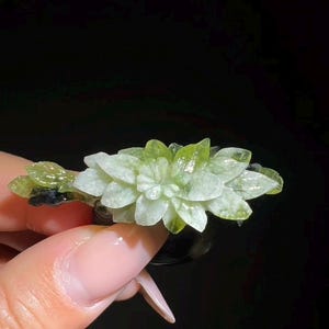 May include: A light green and white floral-shaped crystal. The crystal is held against a black background. The crystal has a detailed, layered design, with a mix of light green and white hues. The overall shape resembles a flower.