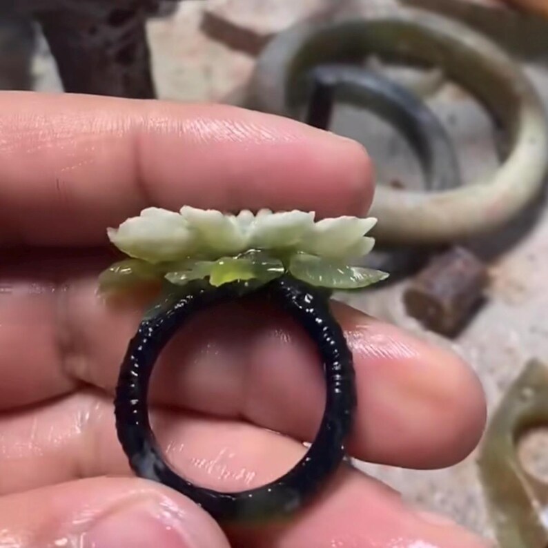 以下が含まれることがあります： 暗いバンドと繊細な花の模様が特徴の手作りの指輪。指輪のバンドは暗く、おそらく黒またはネイビーで、花の要素は明るい緑と白です。指輪は手で持たれており、複雑なディテールが示されています。