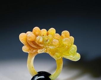 May include: A unique ring featuring a floral design in shades of yellow and orange. The ring's band is a light yellow, and it has a black, rounded base. The flower has a detailed, layered appearance, creating a striking piece of jewelry.